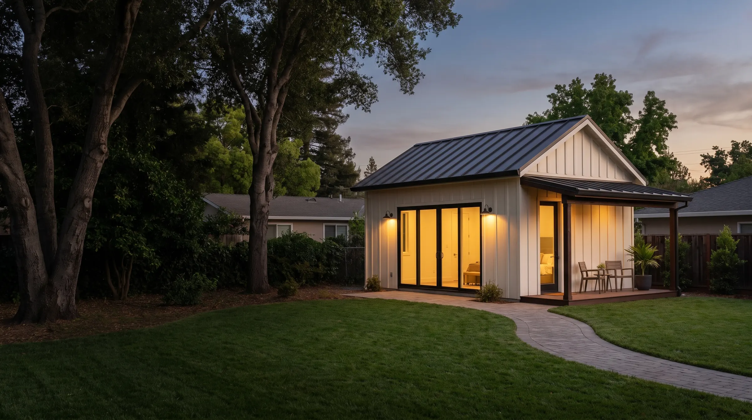 Detached backyard ADU at dusk with white board-and-batten siding, a standing-seam metal roof and glowing glass doors behind a San Jose home